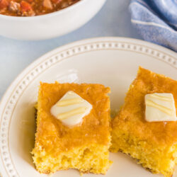 Two pieces of cornbread on a plate with a bowl of chili in the background