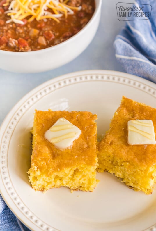 Two pieces of cornbread on a plate with a bowl of chili in the background