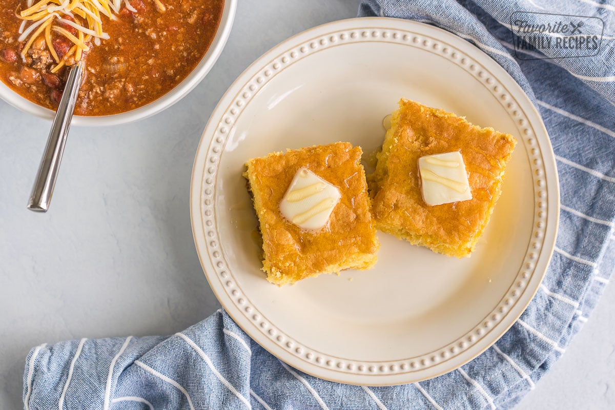 Two pieces of cornbread on a plate with a bowl of chili in the background