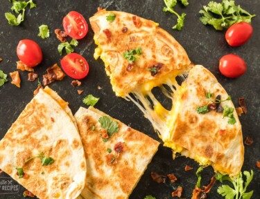 Golden, crispy breakfast quesadillas filled with eggs, cheese, and bacon on a black tabletop.