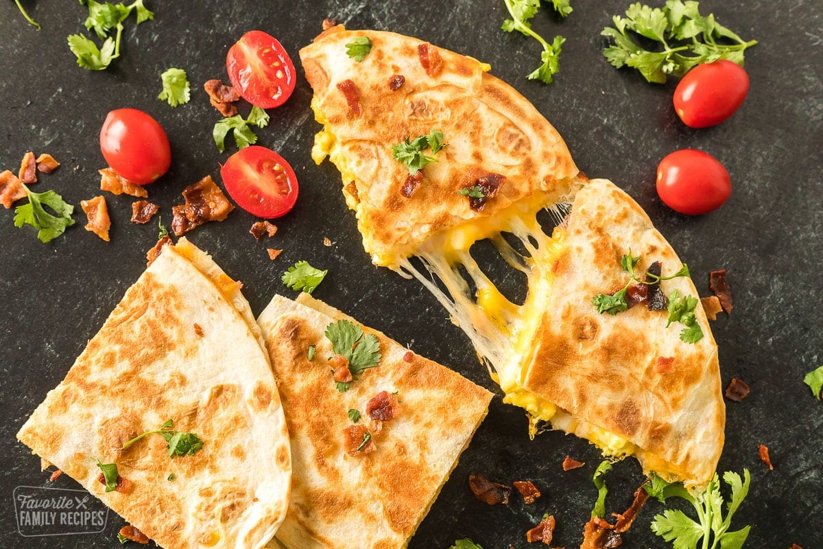 Golden, crispy breakfast quesadillas filled with eggs, cheese, and bacon on a black tabletop.