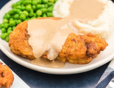 Chicken fried chicken on a plate covered in gravy with mashed potatoes and peas