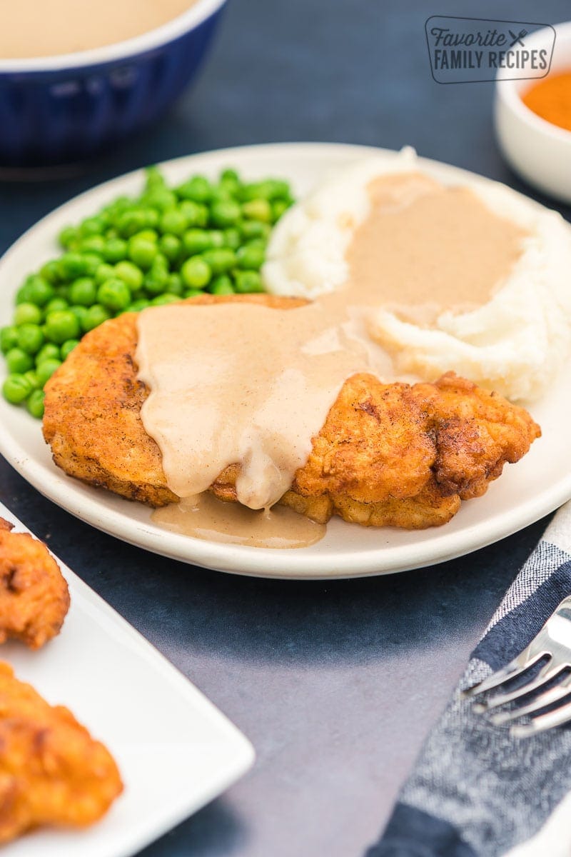 Chicken fried chicken on a plate covered in gravy with mashed potatoes and peas