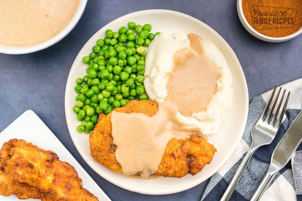 Chicken fried chicken on a plate covered in gravy with mashed potatoes and peas