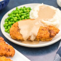 Chicken fried chicken on a plate covered in gravy with mashed potatoes and peas