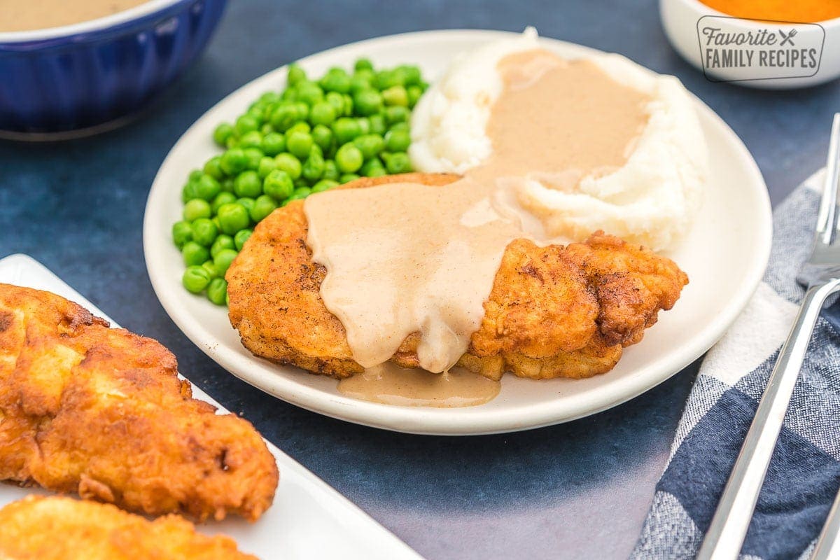 Chicken fried chicken on a plate covered in gravy with mashed potatoes and peas