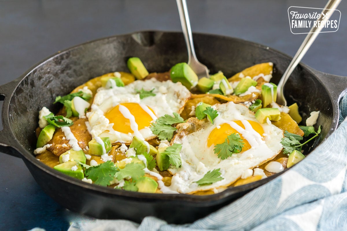 Two forks in a cast iron skillet full of chilaquiles - tortilla chips cooked in salsa verde, eggs, avocado and cilantro.