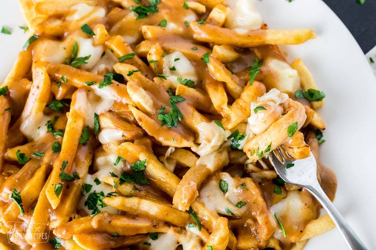 A close up of a plate of fries with gravy and cheese curds (poutine) with a fork lifting a bite.