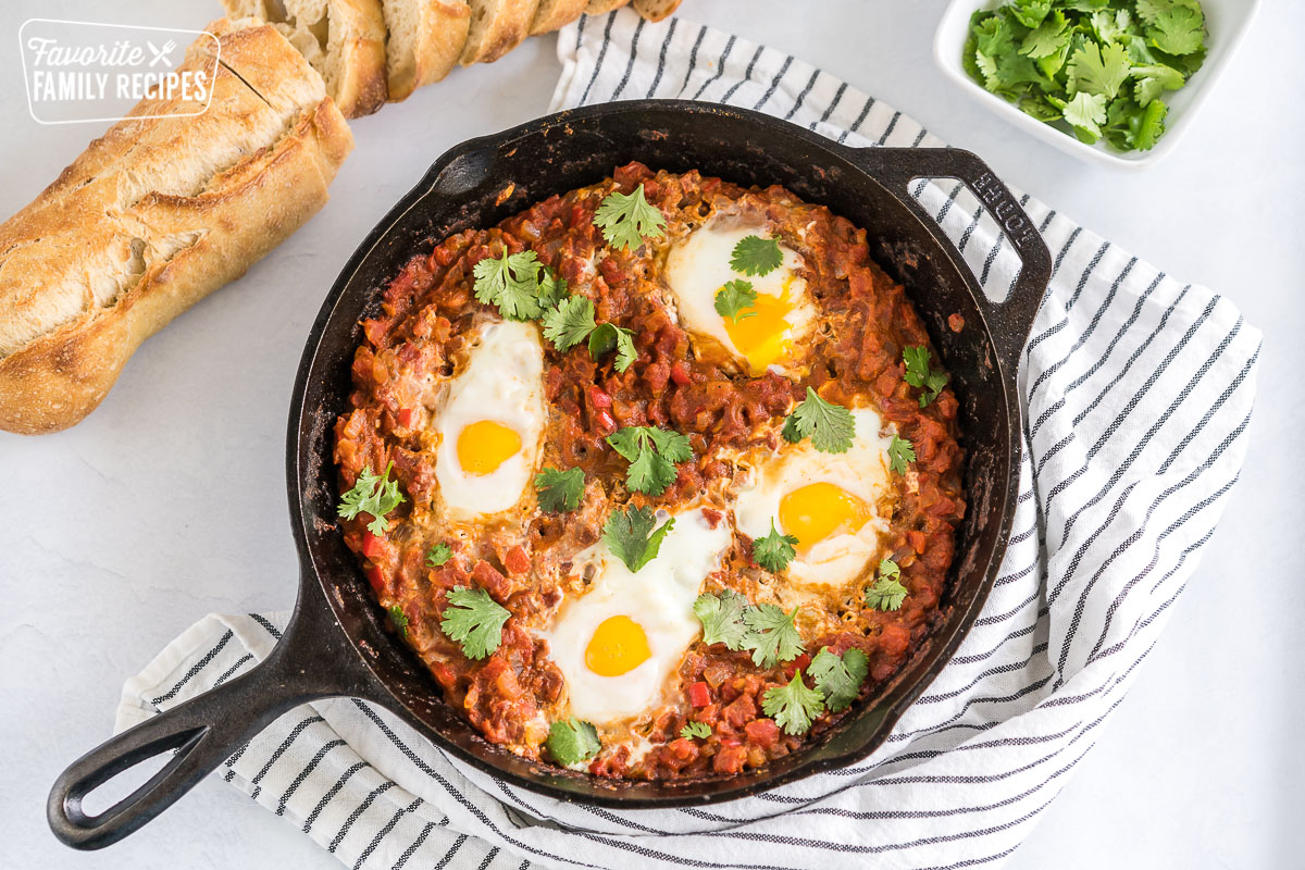 Shakshuka in a cast iron pan with a loaf of bread and a bowl of cilantro.