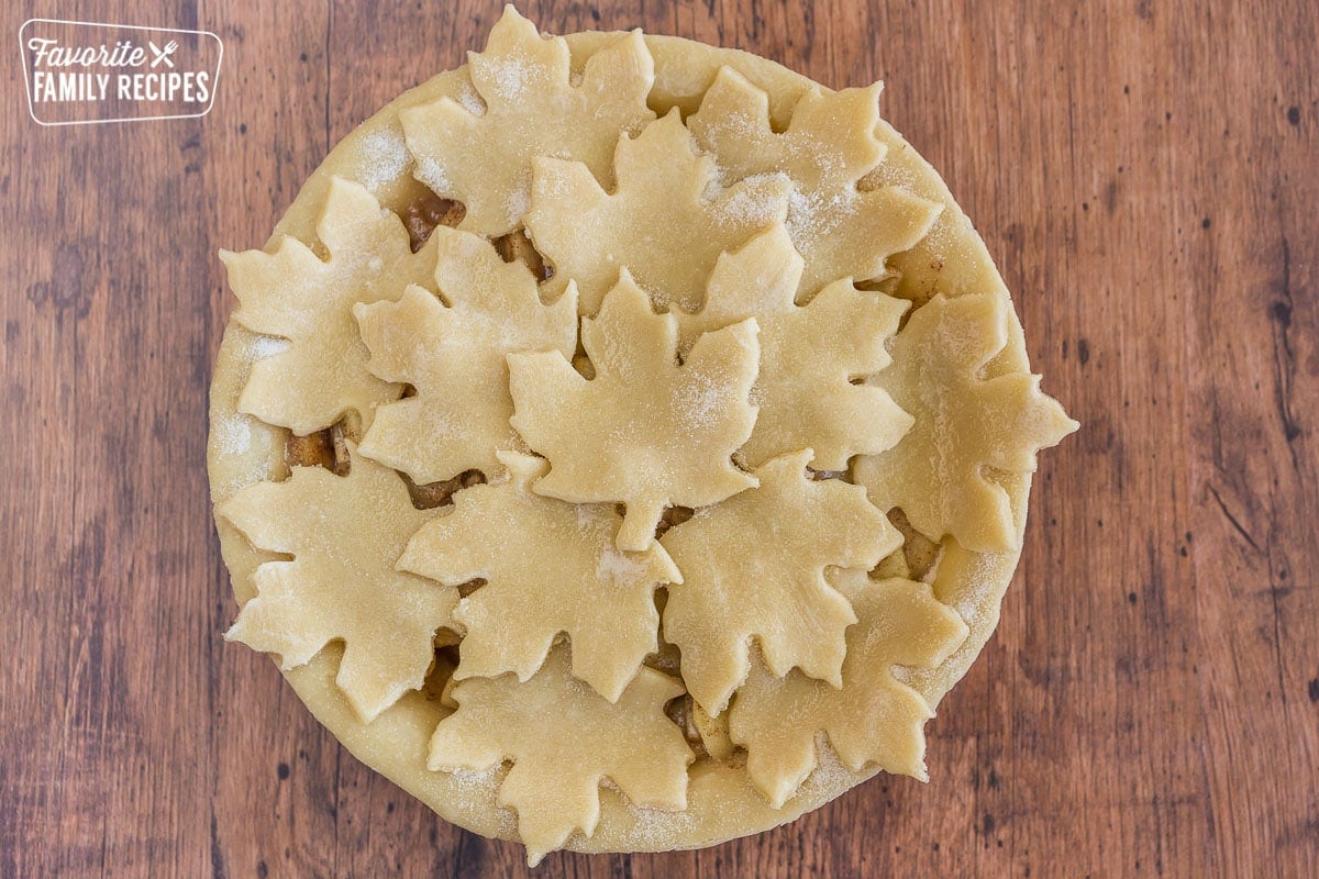 An uncooked apple pie topped with pie crust dough cut outs shaped like maple leaves on top and milk and sugar