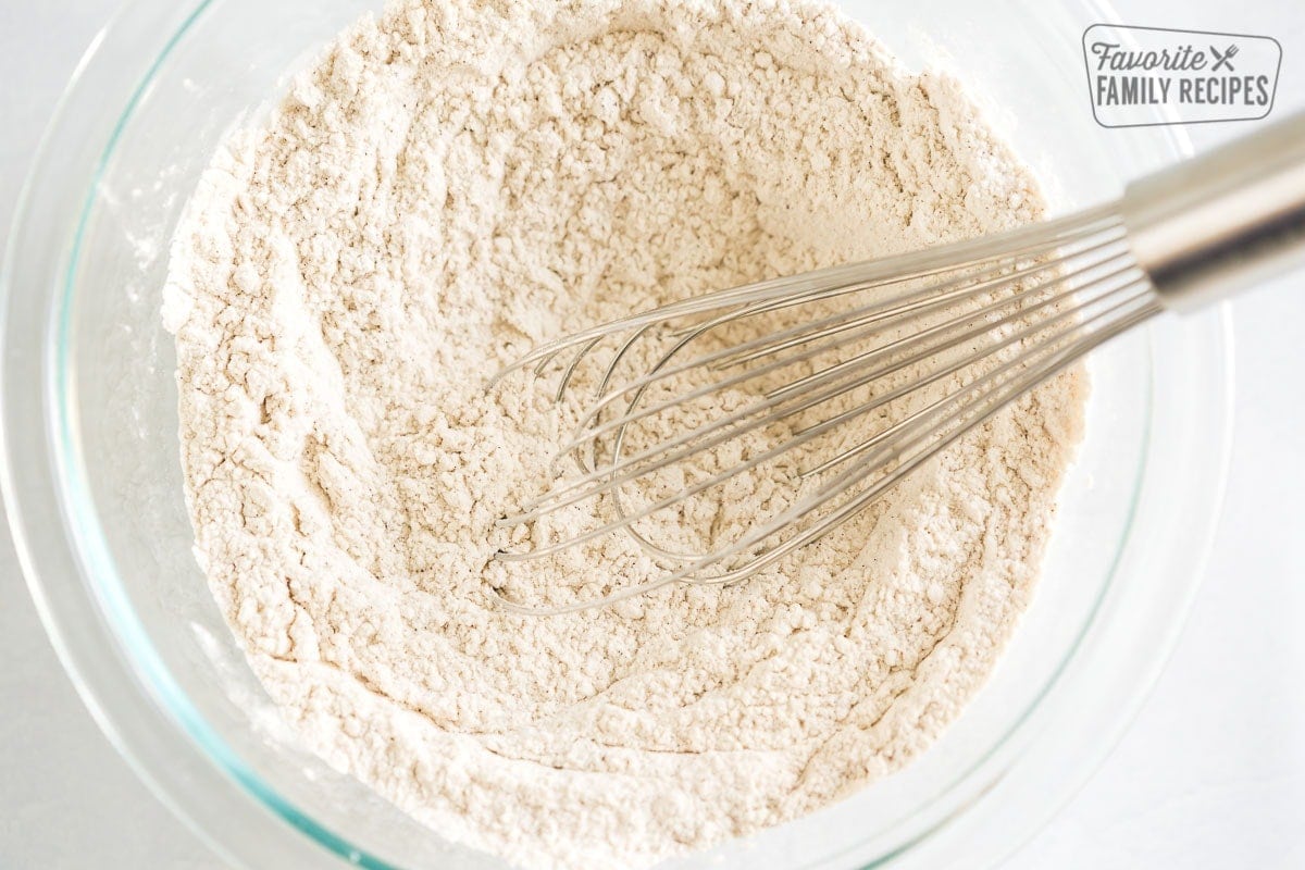 A glass bowl with flour, baking soda, nutmeg, cinnamon, and salt being stirred with a whisk