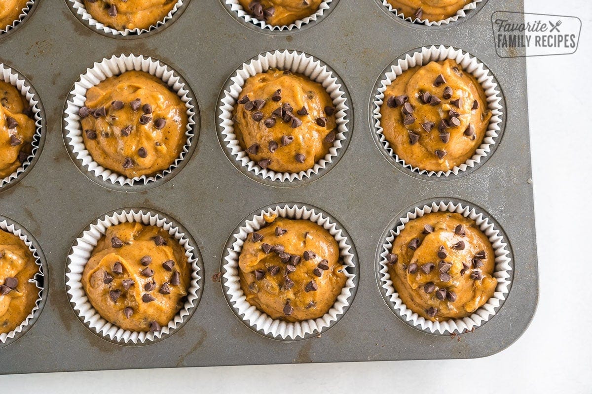 A muffin tin with white liners filled with batter and topped with chocolate