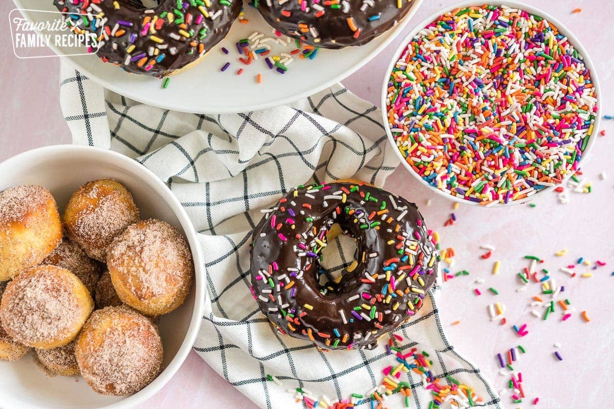 A chocolate sprinkle donut next to a bowl of sprinkles and a bowl of donut holes