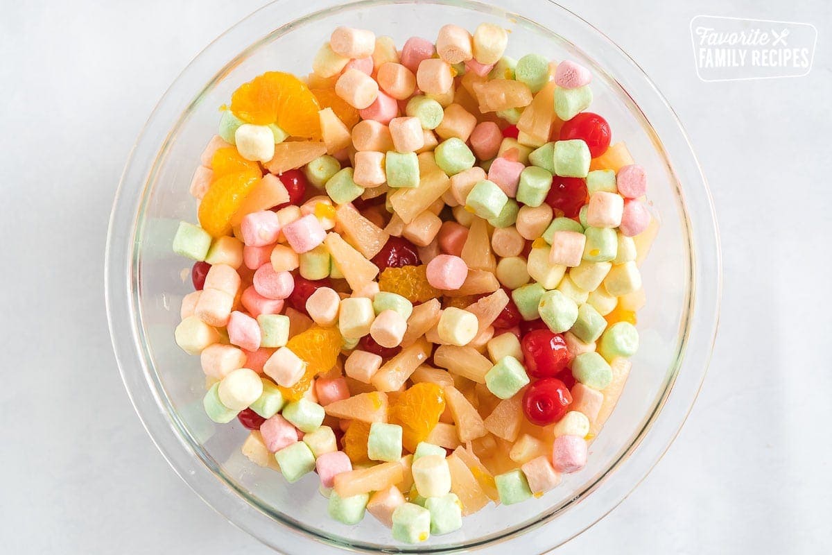 A glass bowl full of marshmallows, mandarin oranges, pineapple, and cherries