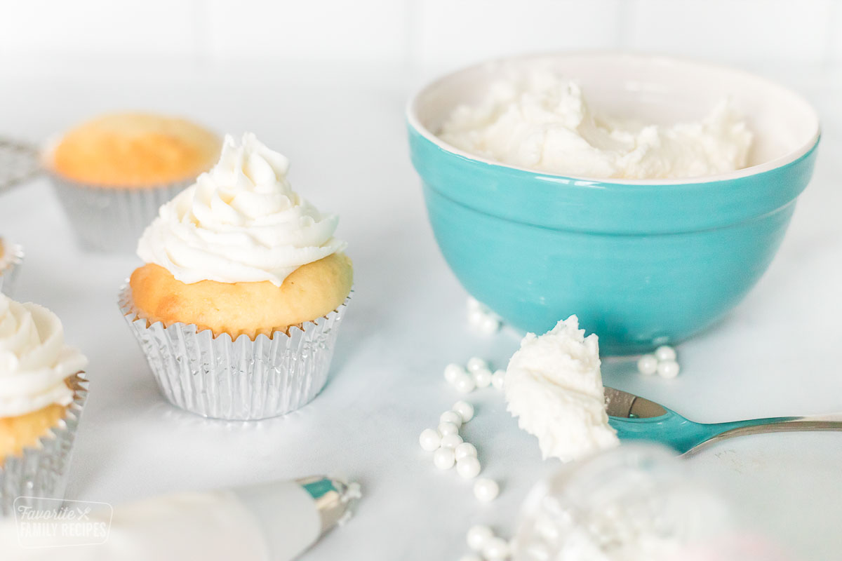 A cupcake next to a bowl of buttercream frosting and a spoon with a scoop of frosting on it