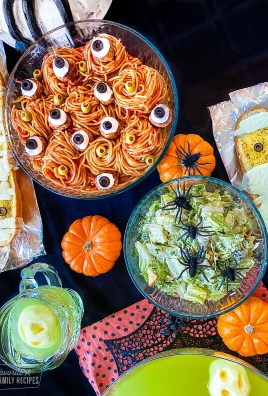 Spaghetti with meatballs that have cheese and olives that look like eyes, green punch, French bread with cheese, and a Caesar salad with plastic spiders for a Halloween dinner.