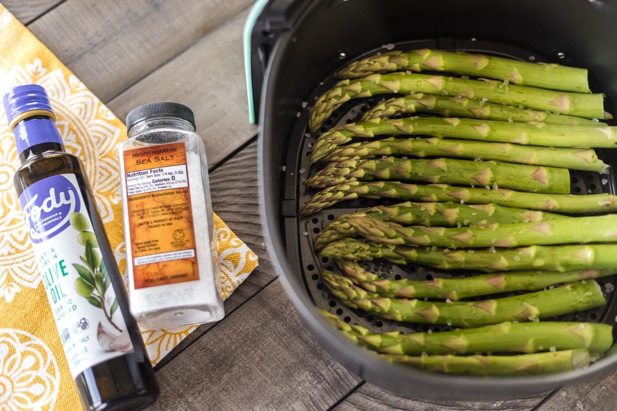 Asparagus in an air fryer next to a bottle of olive oil and a jar of salt