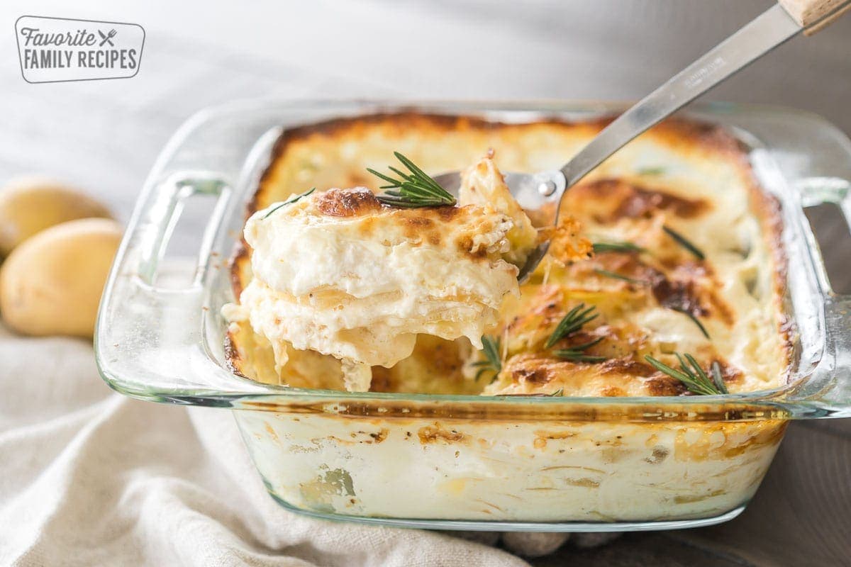 A scoop of Au Gratin potatoes being taken out of a baking dish with a large metal serving spoon