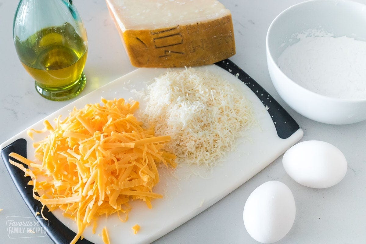 Shredded cheddar and parmesan cheese on a cutting board next to two eggs and a bottle of oil