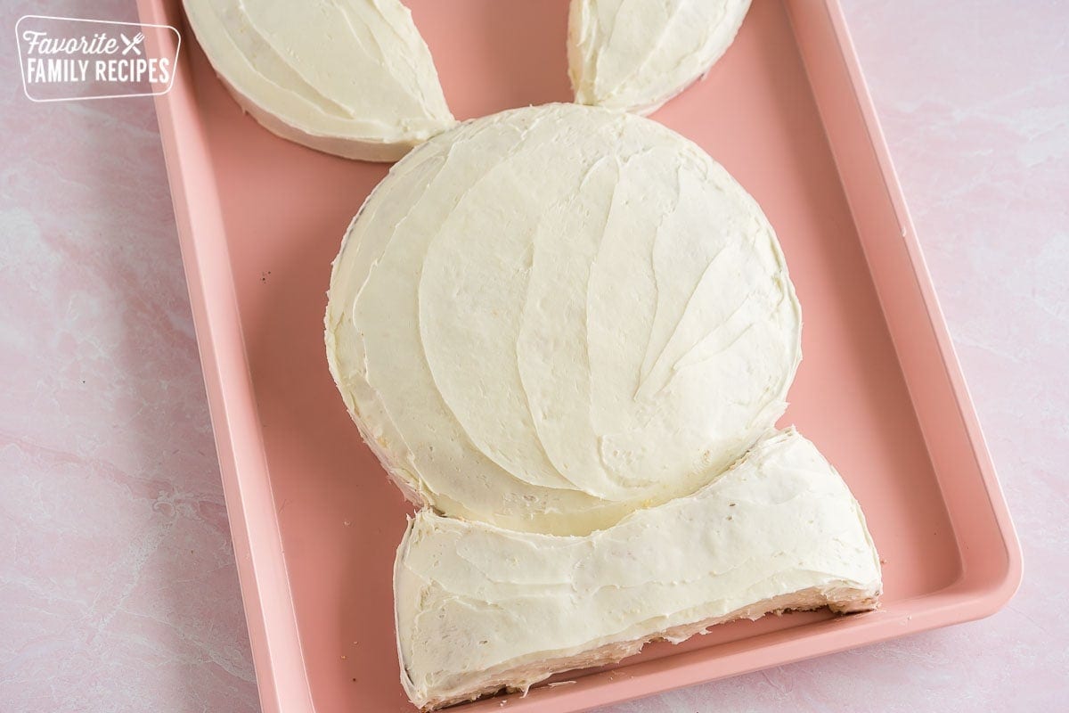 A cake in the shape of a bunny frosted with white frosting on a pink baking sheet
