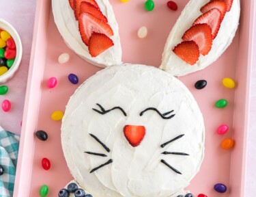 A bunny cake on a pink baking sheet decorated with strawberries, blueberries, and black icing in the shape of eyes and whiskers.