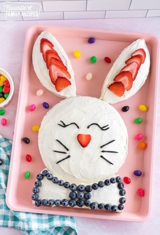 A bunny cake on a pink baking sheet decorated with strawberries, blueberries, and black icing in the shape of eyes and whiskers.