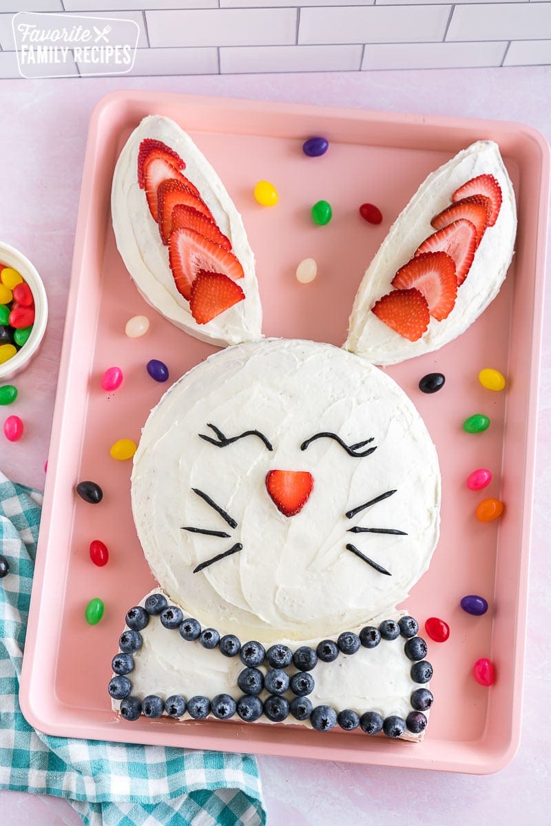 A bunny cake on a pink baking sheet decorated with strawberries, blueberries, and black icing in the shape of eyes and whiskers.