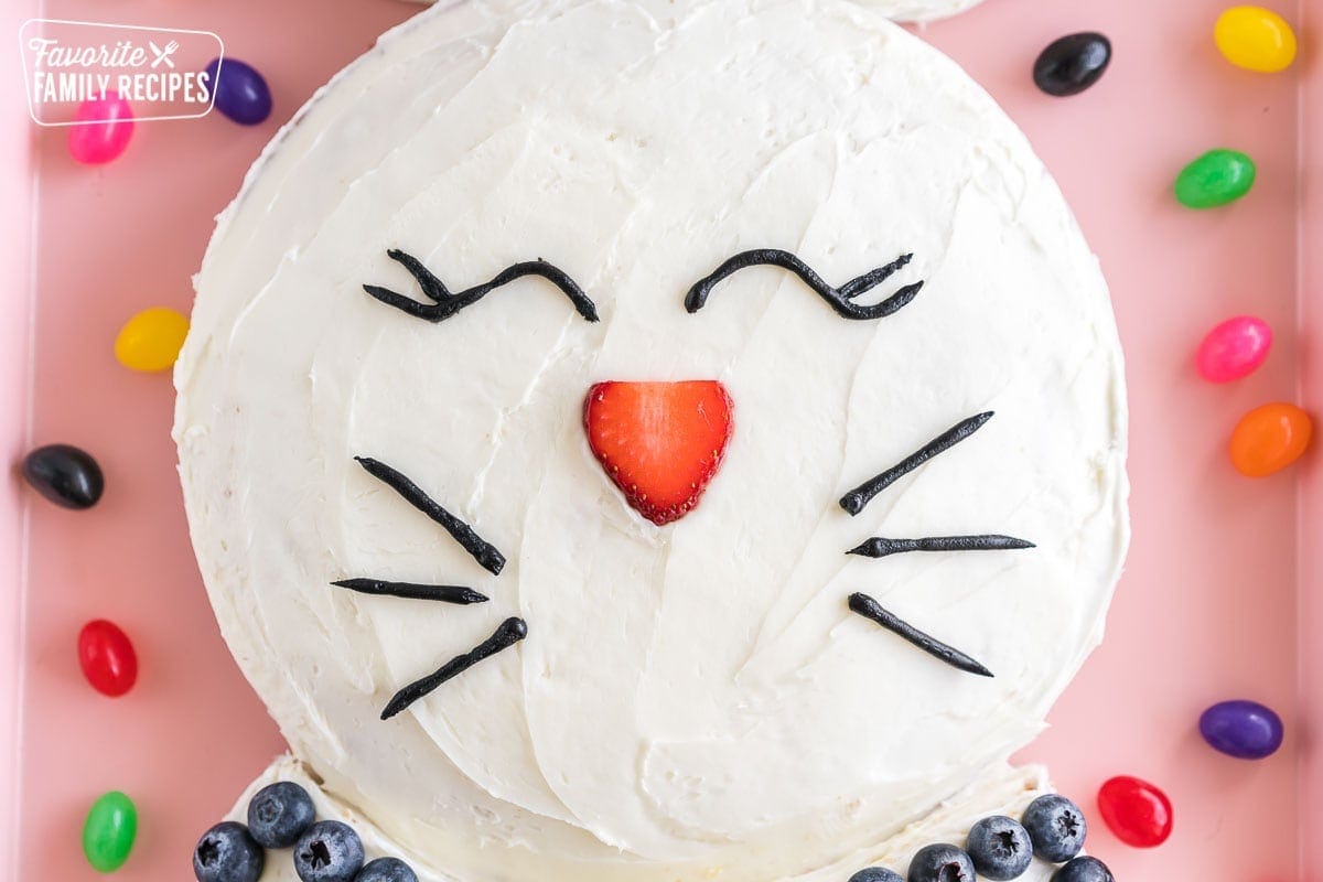 A round cake frosted with white frosting and decorated with a strawberry slice as a nose and black icing eyes and whiskers
