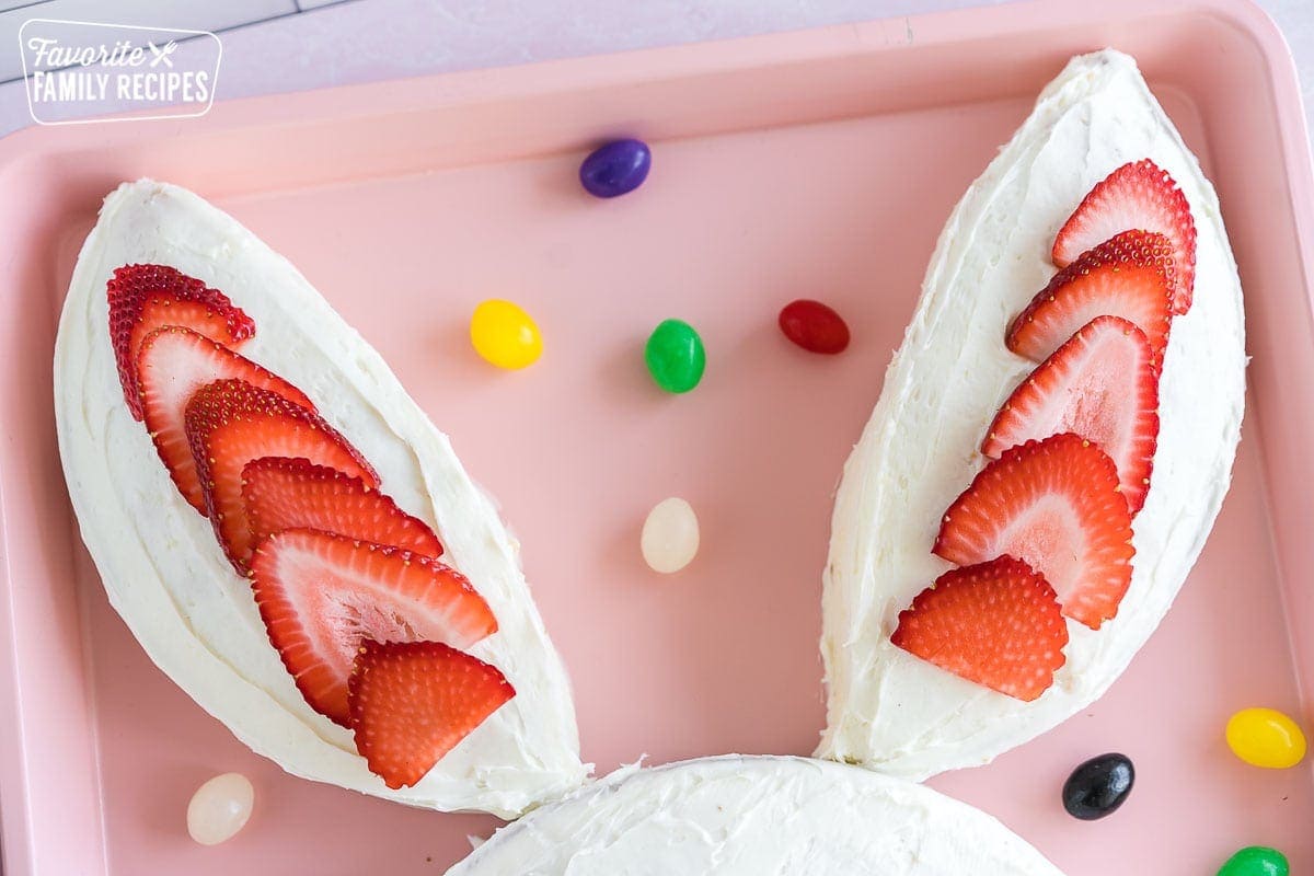 Cake shaped like two bunny ears, frosted with white frosting and decorated with strawberry slices