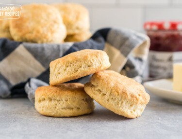 Three buttermilk biscuits stacked on each other with a stick of butter and a jar of jam in the background