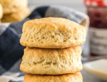 Three buttermilk biscuits stacked on each other with a stick of butter and a jar of jam in the background