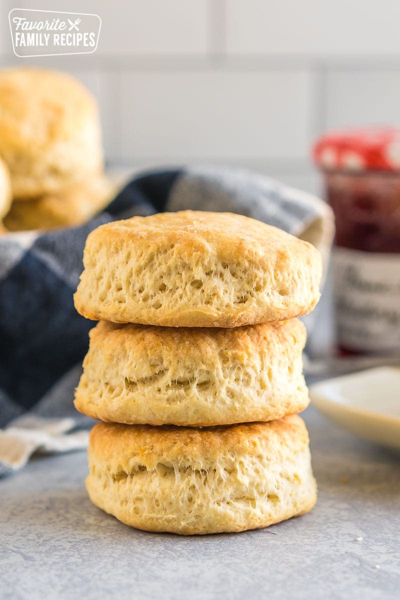Three buttermilk biscuits stacked on each other with a stick of butter and a jar of jam in the background