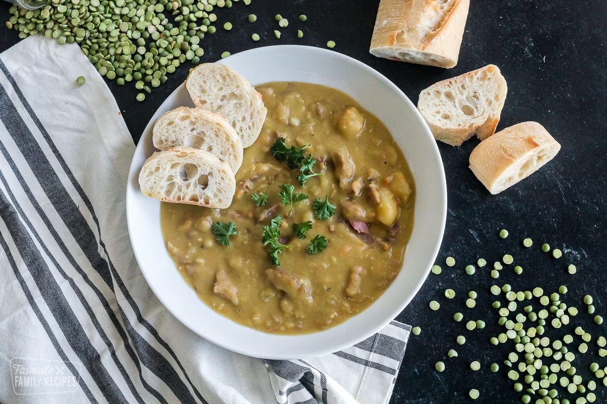 A bowl of CrockPot split pea soup next to slices of bread and uncooked split peas.