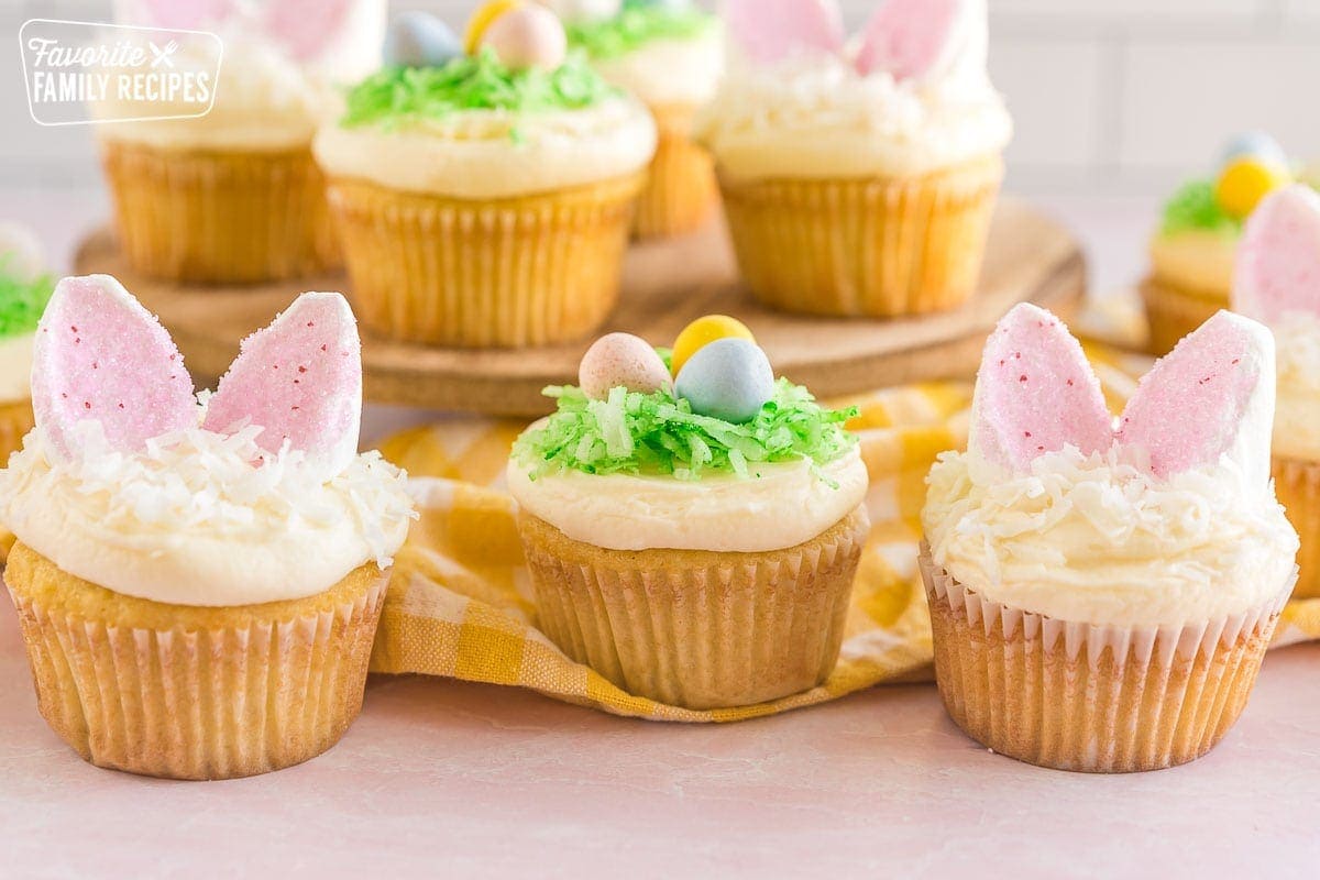 Three Easter Cupcakes, one with a green coconut birds nest with candy eggs and two with marshmallow bunny ears