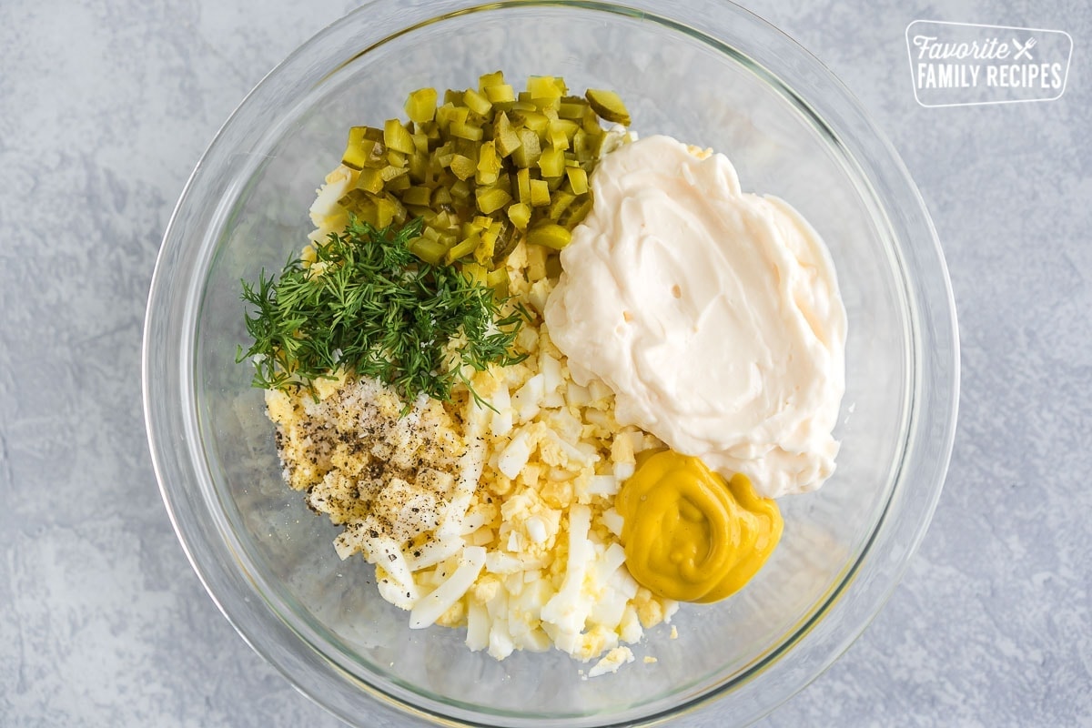A large glass bowl containing sliced egg, salt, pepper, chopped pickles, chopped dill, mayonnaise, and mustard.