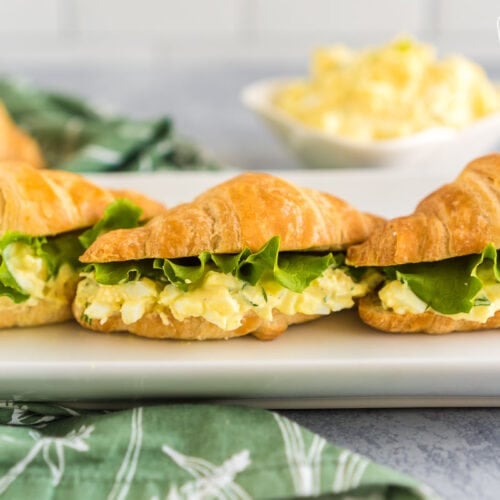 Three Egg Salad Sandwiches on a white platter. Each egg salad sandwich is made on a croissant with a slice of lettuce on top