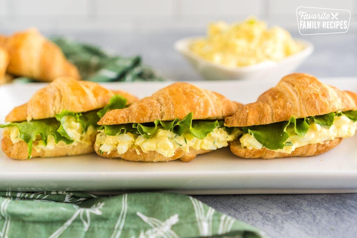 Three Egg Salad Sandwiches on a white platter. Each egg salad sandwich is made on a croissant with a slice of lettuce on top.