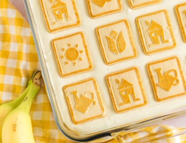 A baking dish of banana pudding with a yellow gingham towel and two bananas