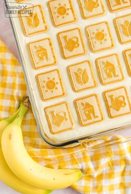 A baking dish of banana pudding with a yellow gingham towel and two bananas