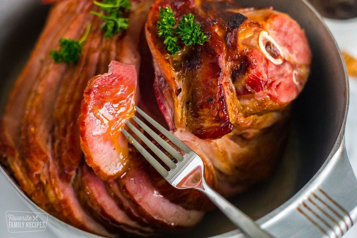 A whole ham in a roasting pan with a slice being removed with a fork
