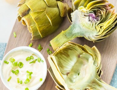 Cooked artichoke halves on a cutting board with dipping sauce