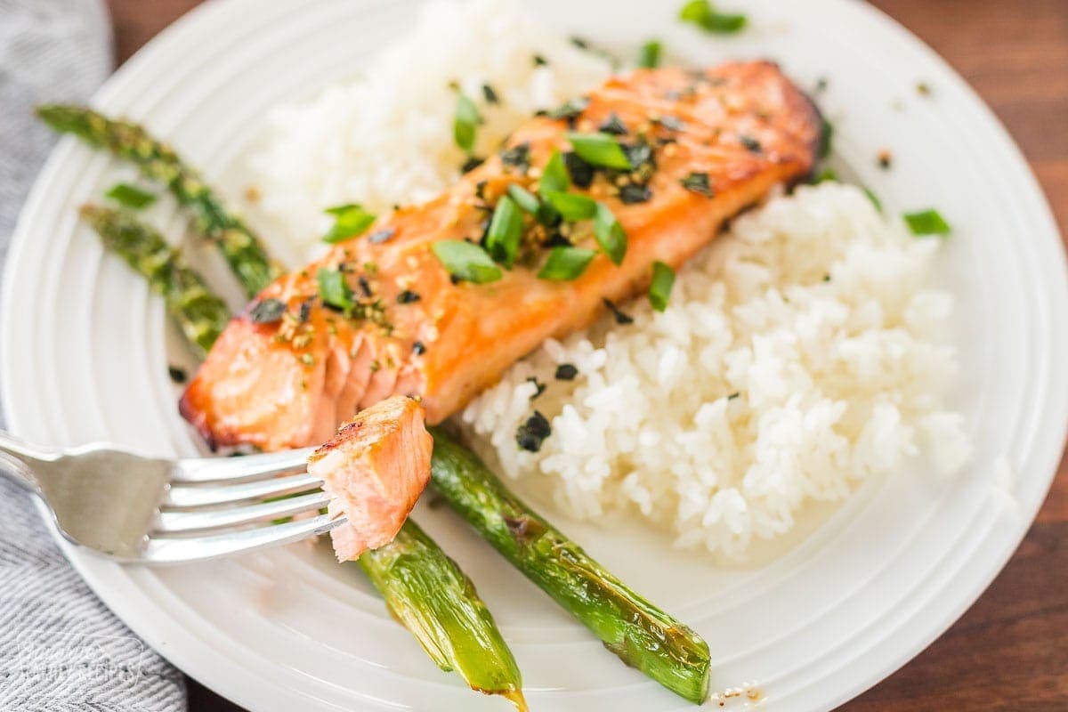 A close up of a fork with a piece of miso salmon on it. A plate of salmon with rice and asparagus is in the background