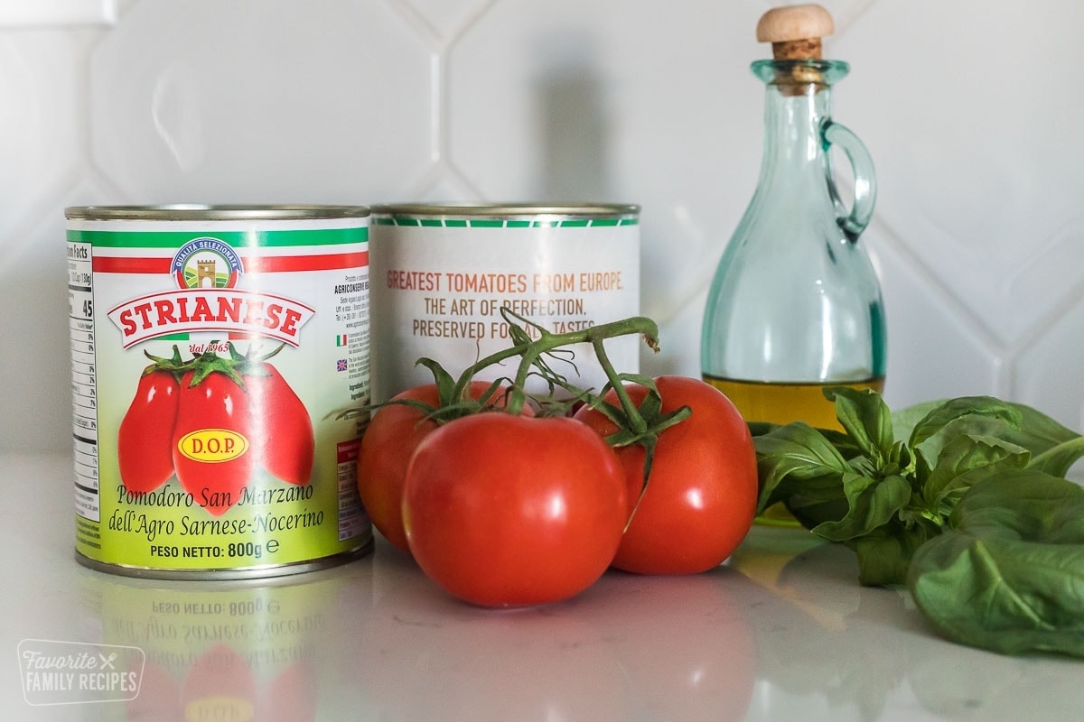 Two cans of San mMrzano tomatoes, three tomatoes, a jar of olive oil, and some fresh basil on a counter
