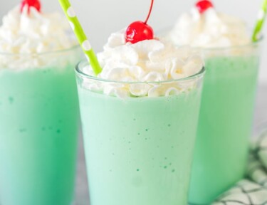 A mint green Shamrock shake in a clear glass topped with whipped cream, a maraschino cherry, and a straw.