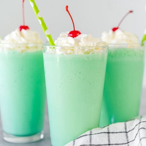 A mint green Shamrock shake in a clear glass topped with whipped cream, a maraschino cherry, and a straw.