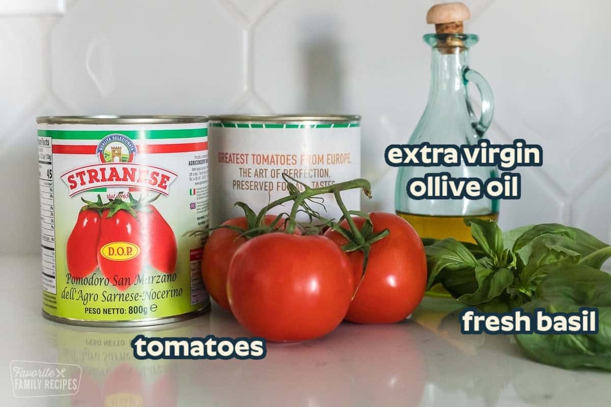 Two cans of San Marzano tomatoes, three tomatoes, a jar of olive oil, and some fresh basil on a counter