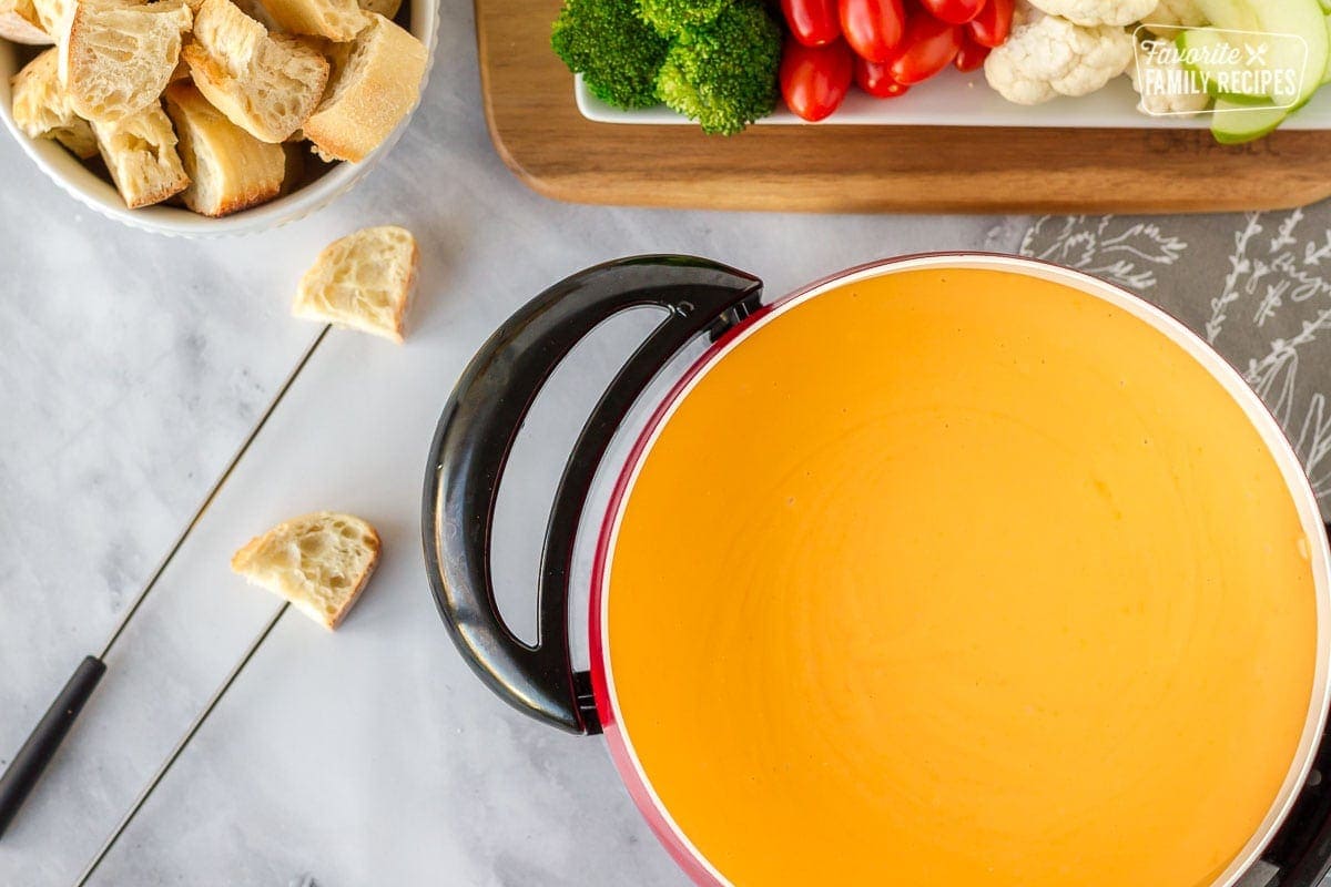Cheddar fondue in a fondue pot with sliced bread in skewers and a bowl with extra sliced bread and vegetables on a dish for dipping.