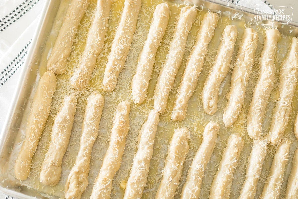 Uncooked and un-risen breadsticks sitting in melted butter on a jelly roll pan with parmesan cheese sprinkled over the top