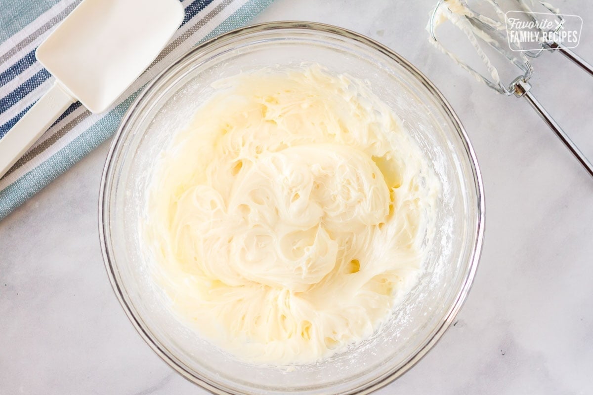 Bowl of whipped cream cheese frosting with beaters and spatula on the side used to spread on easy fruit pizza.