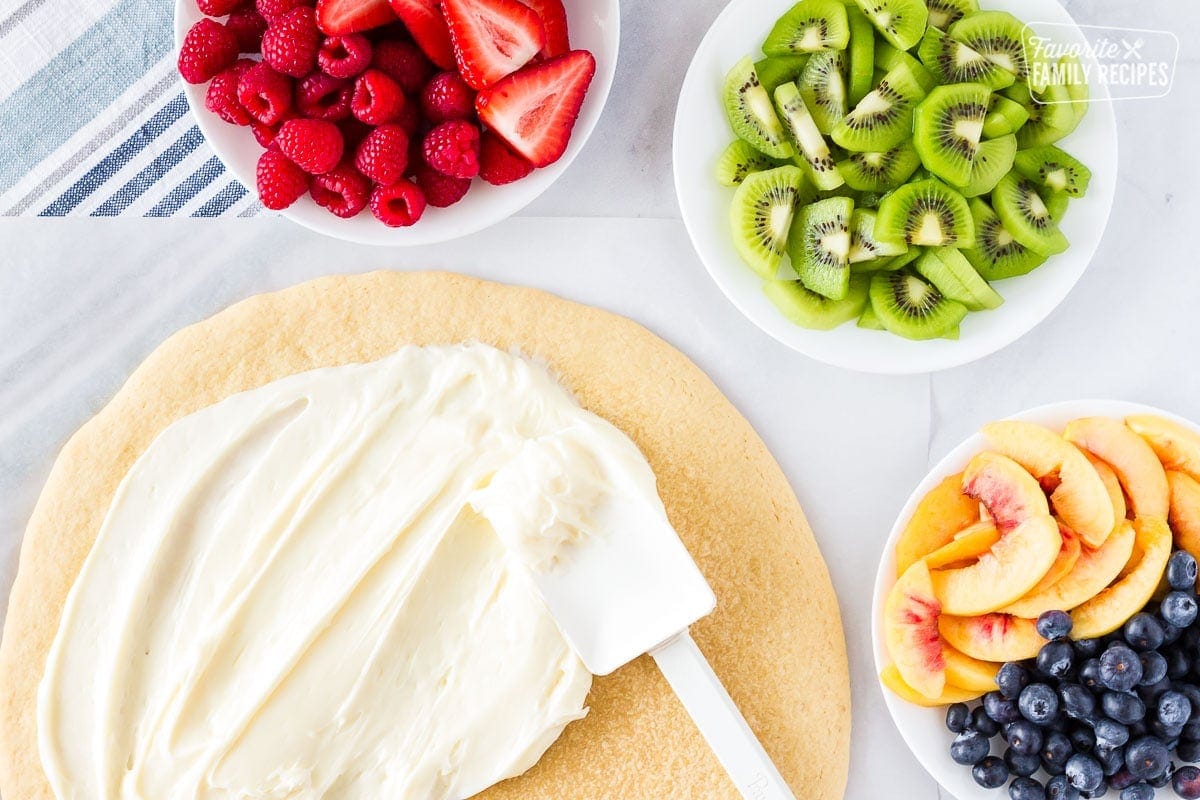 Cookie pizza with a spatula spreading cream cheese frosting and three bowls of fresh fruit on the side.